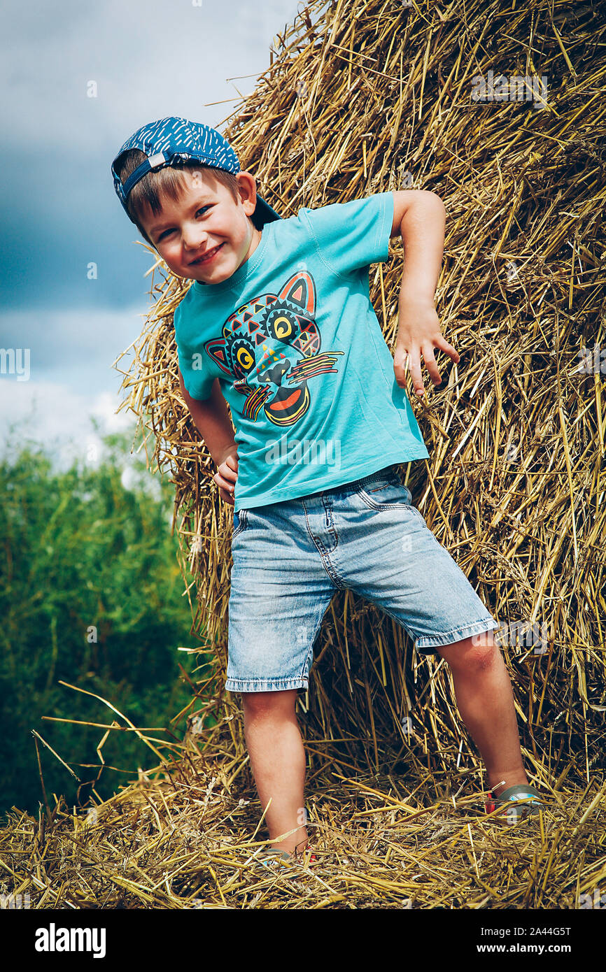A boy in a blue baseball cap and shorts having fun and playing on round bales of straw on a hot summer day Stock Photo