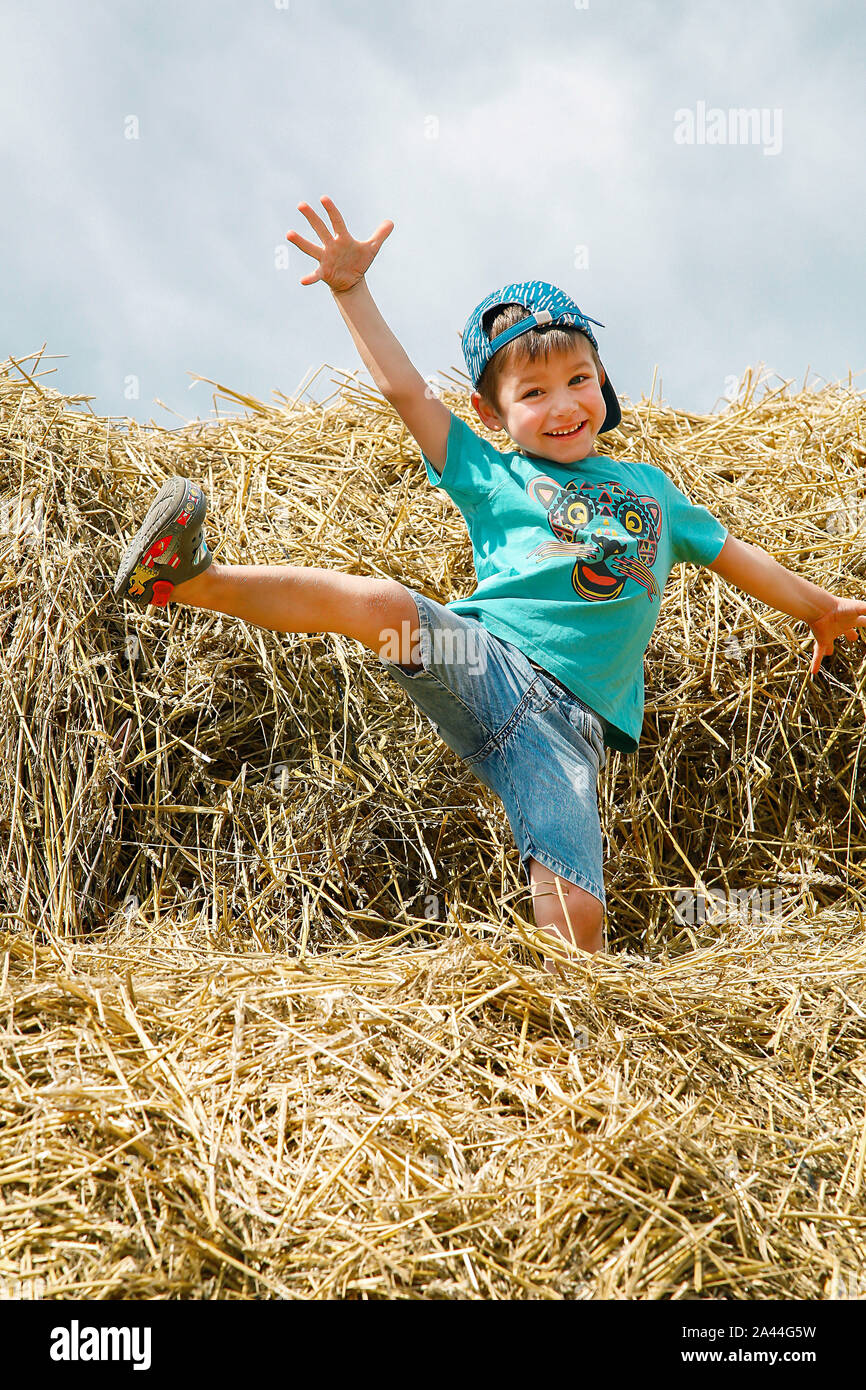 A schoolboy in a blue baseball cap and short shorts plays and has fun on straw bales on a hot summer day Stock Photo