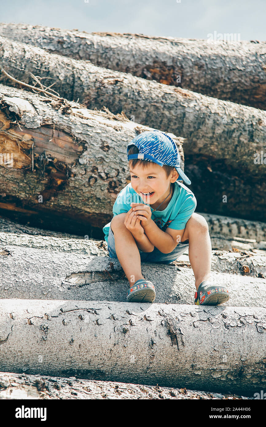 A little boy in a blue baseball cap and short shorts sits on a log and