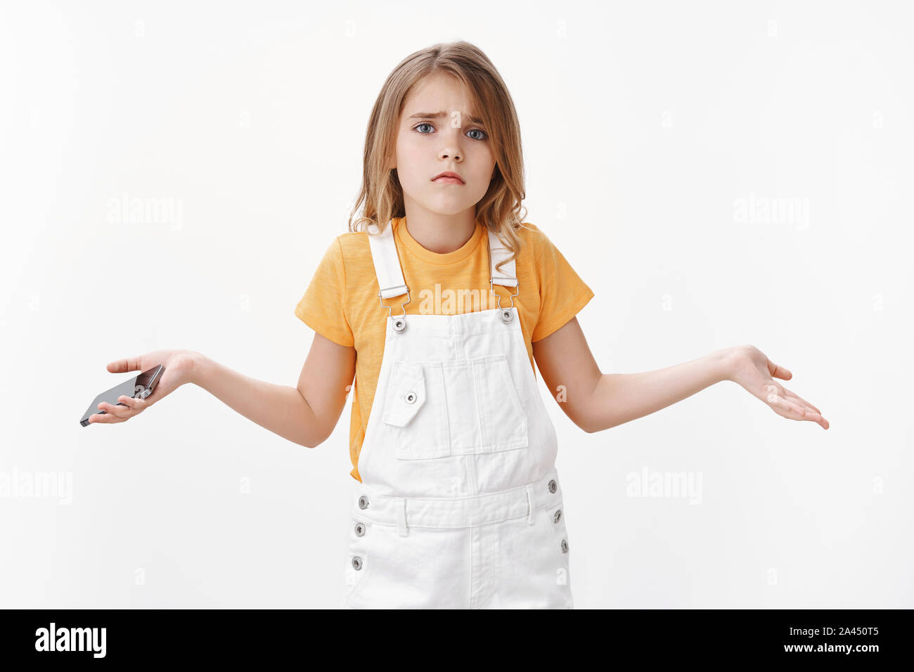Hopeless cute upset little child, girl spread hands sideways desperate, froning upset and disappointed, hold smartphone, losing level in mobile game, Stock Photo