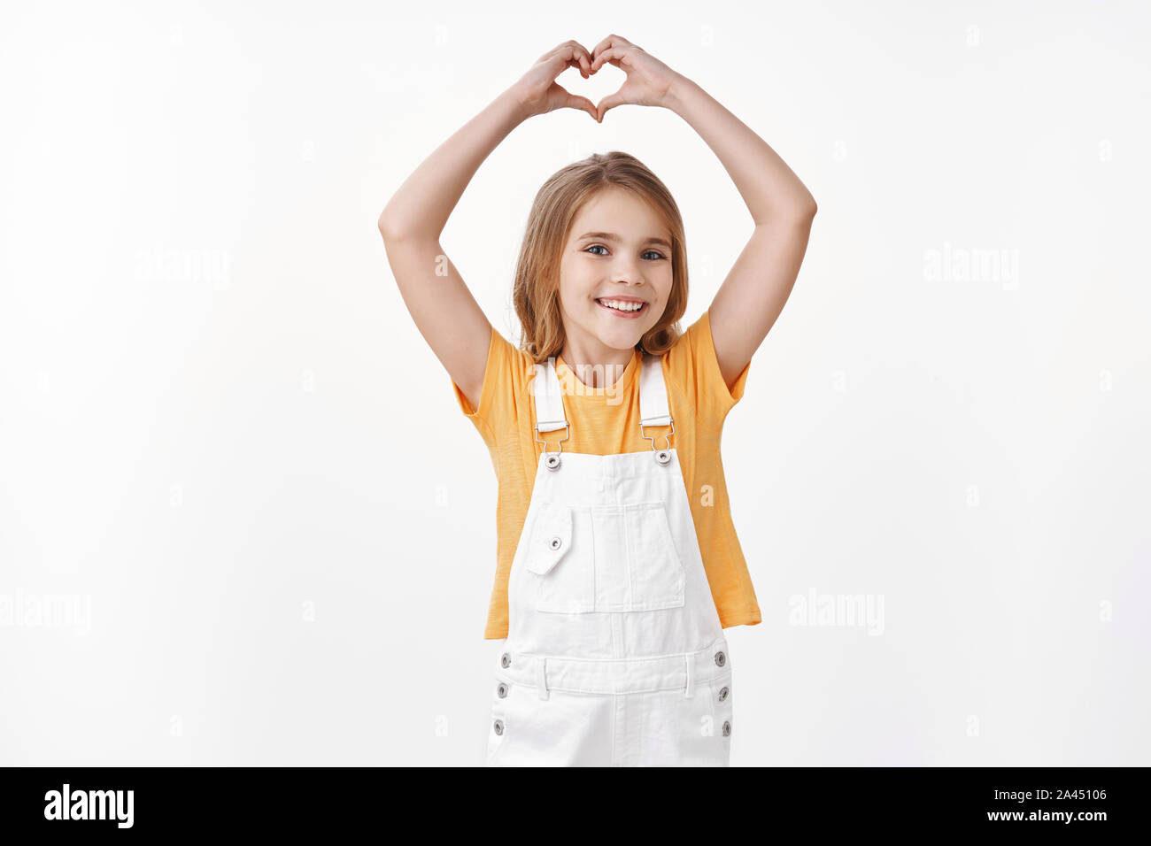 Lovely tender friendly young girl with blond hair, wear summer yellow  t-shirt, overalls, raise hands up show heart sign, confess love to parents,  cher Stock Photo - Alamy