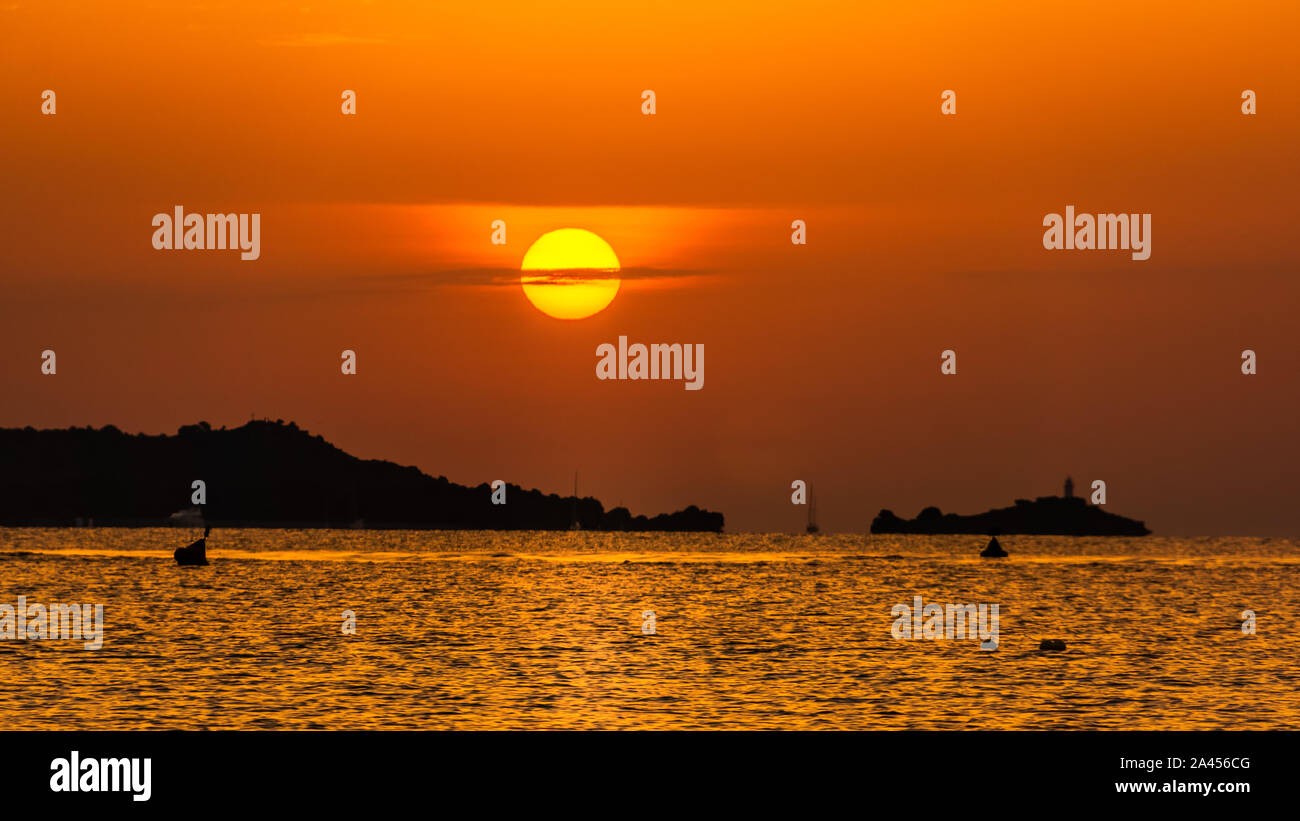 Defined orange solar disc in sunrise with silhouette of mountains, bay of water, reach orange and yellow colours of the sky, panorama, Mallorca, Spain Stock Photo