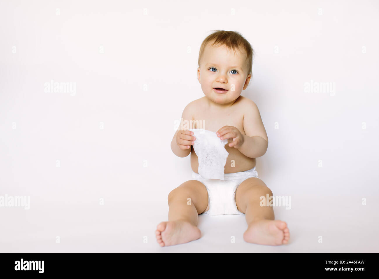 Charming happy little baby girl in white diaper holding wet wipe in her hands and sitting isolated on white background. Cleaning wipe, pure, clean. Stock Photo