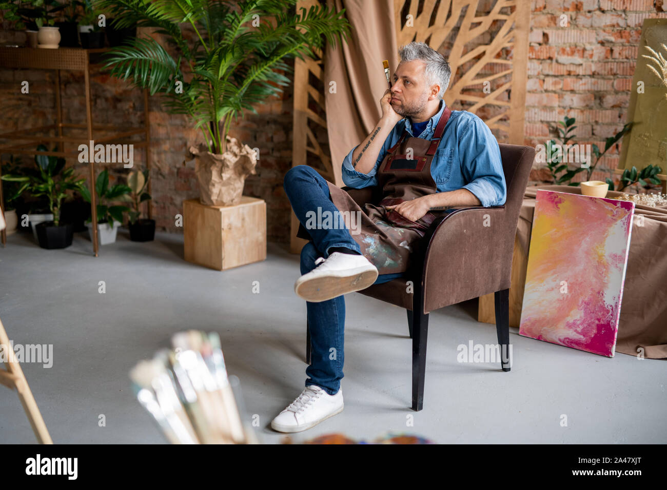 Pensive artist sitting in front of easel and looking at unfinished painting Stock Photo