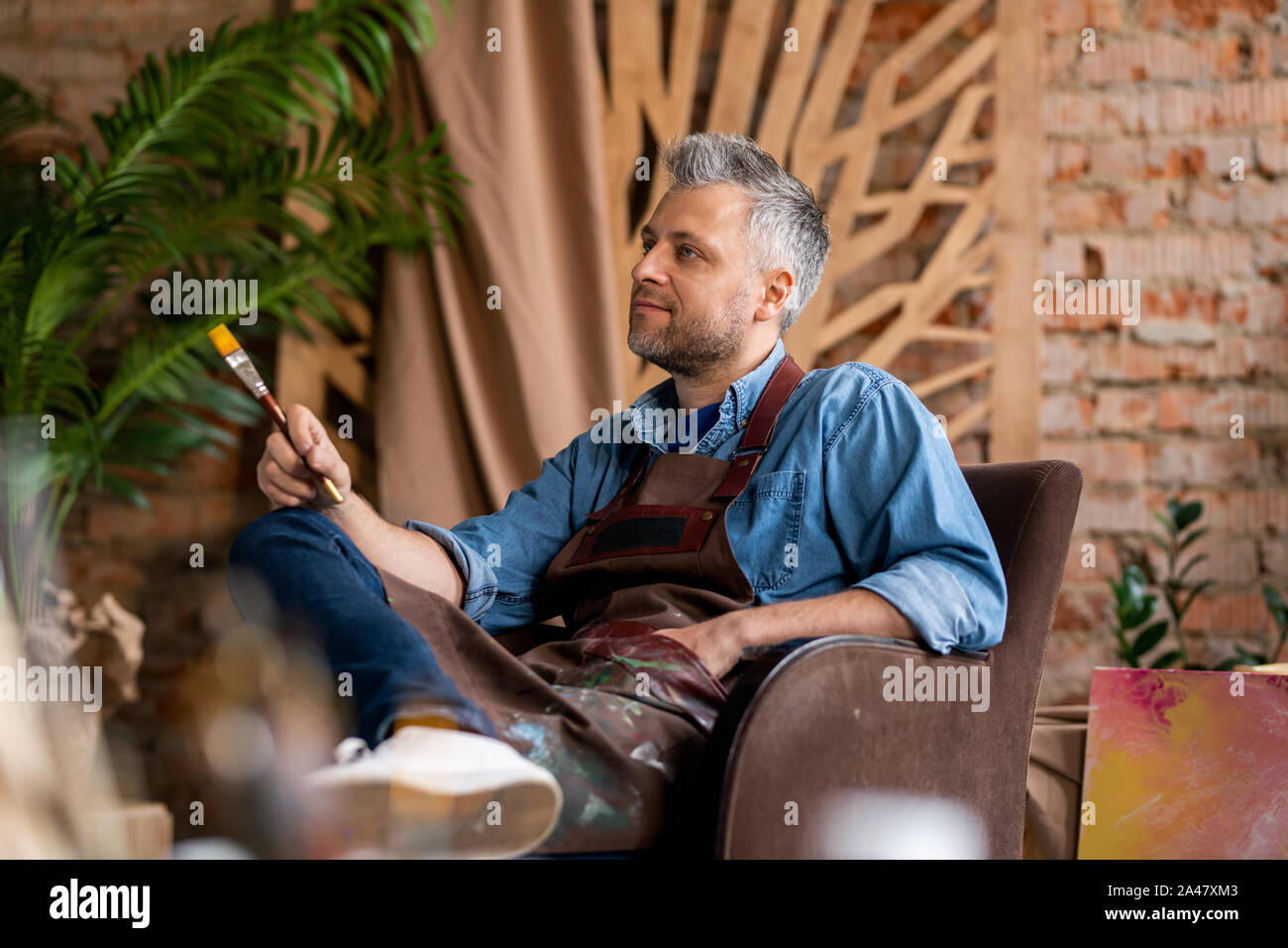 Grey-haired pensive painter with paintbrush sitting in armchair in studio Stock Photo