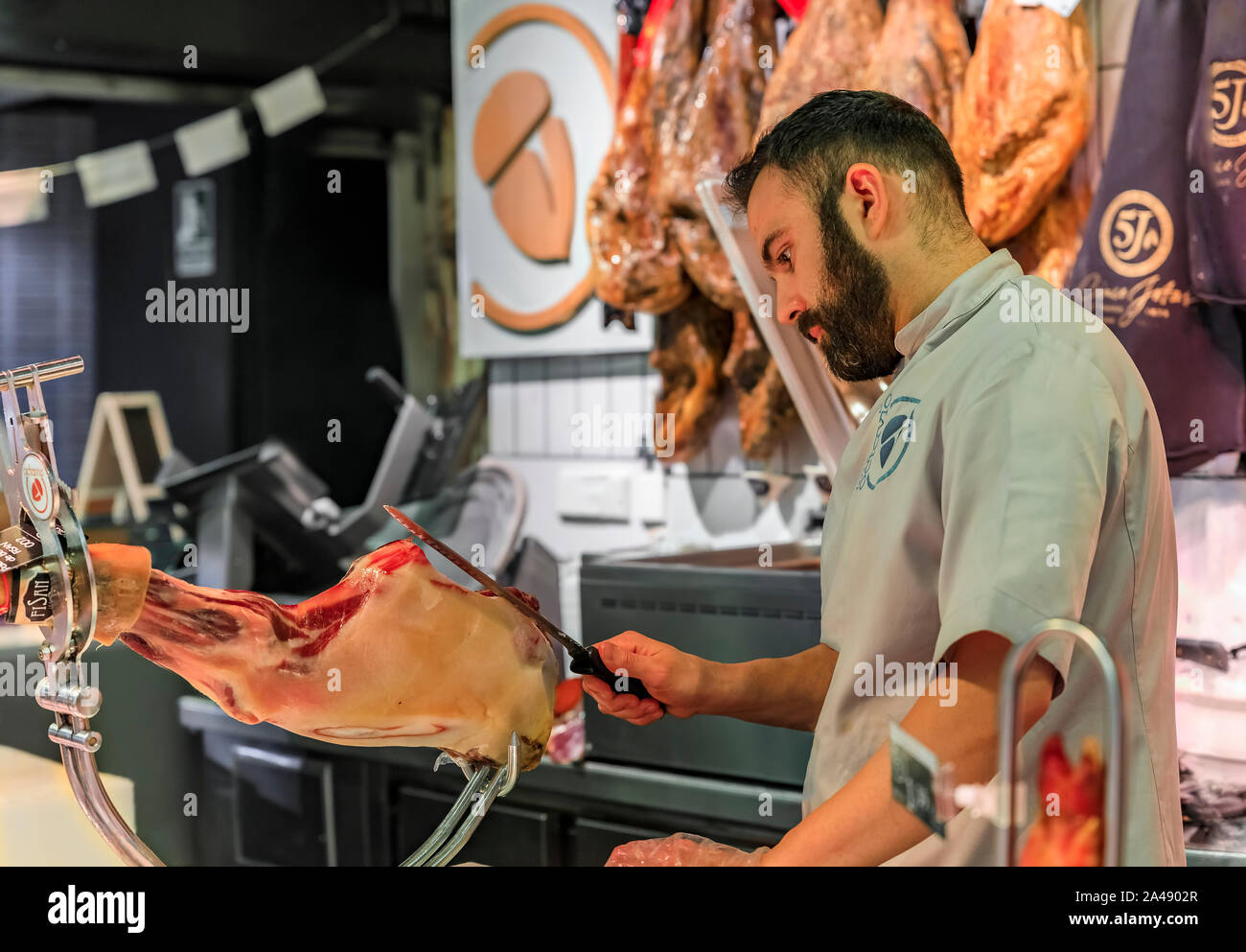 Madrid, Spain - June 4, 2017: Butcher slicing a whole leg of serrano iberico ham on a stand for customers at a market, hams hanging in the background Stock Photo