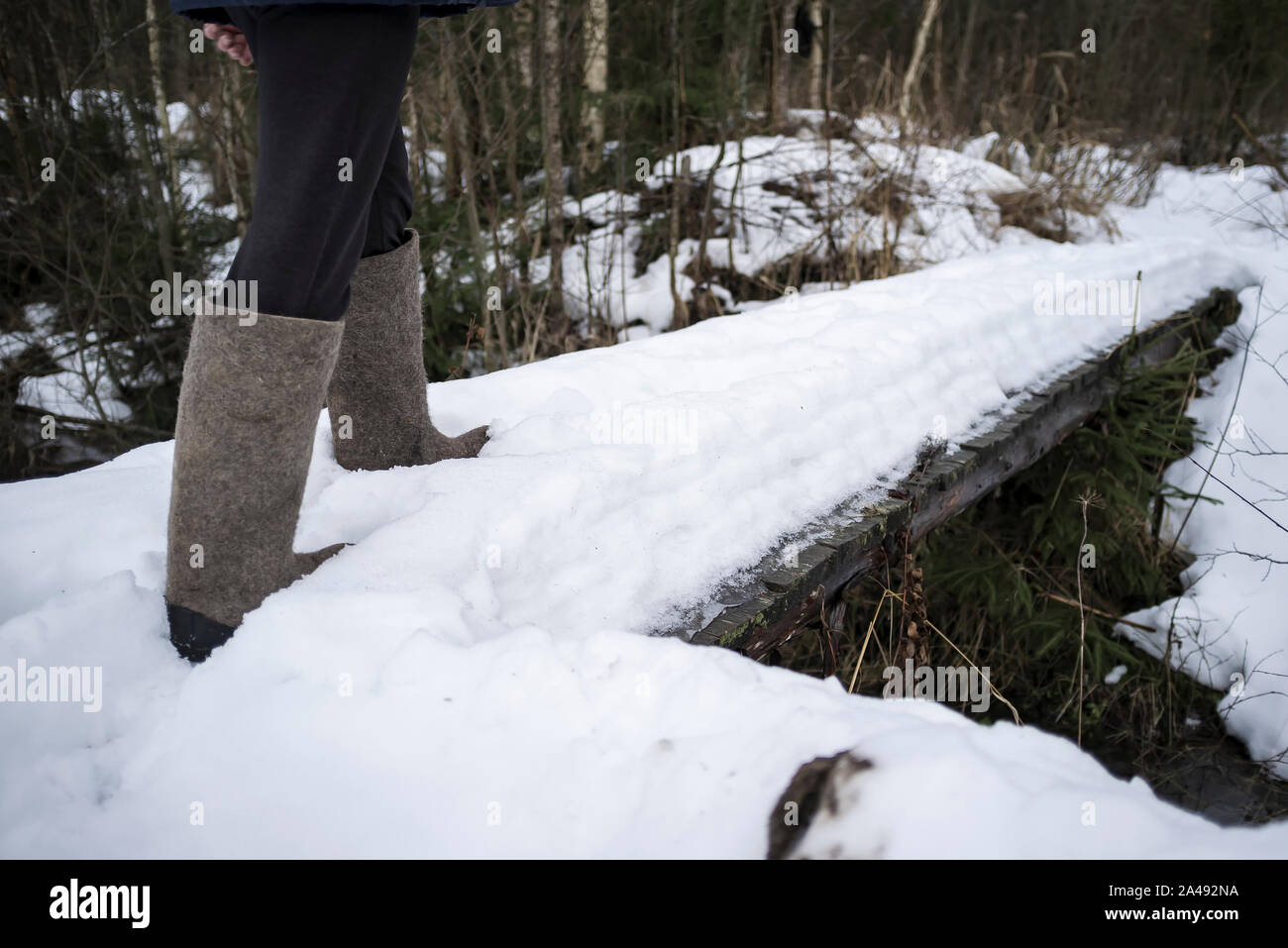 A man walks in a warm traditional winter felt shoes over a small, wooden, snow-covered bridge in the Russian village, on a frosty evening. Close-up. Stock Photo