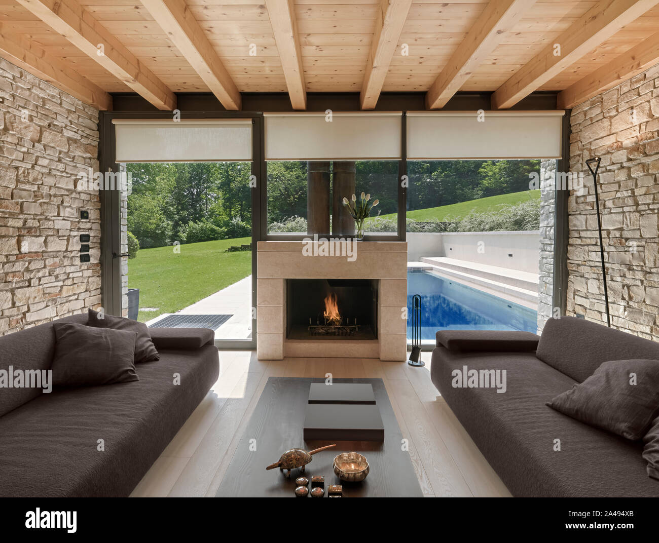 Interior view of a  modern living room whose wall are coated of stone and have a glass wall with a fireplace inteegrated on its, overlooking on the sw Stock Photo