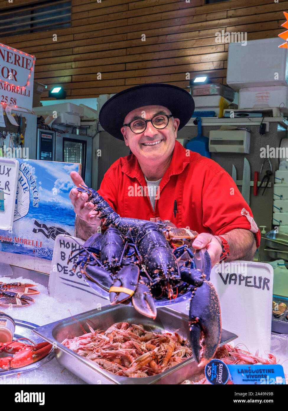 BRETON FISHMONGER & LIVE BLUE LOBSTER HOMARD BLEU FISH MARKET STALL CONCARNEAU FRANCE BRITTANY  Concarneau daily interior French Fish Market hall and character fishmonger, in Breton style outfit proudly displaying his local live blue lobster for sale Brittany Finistere France Stock Photo