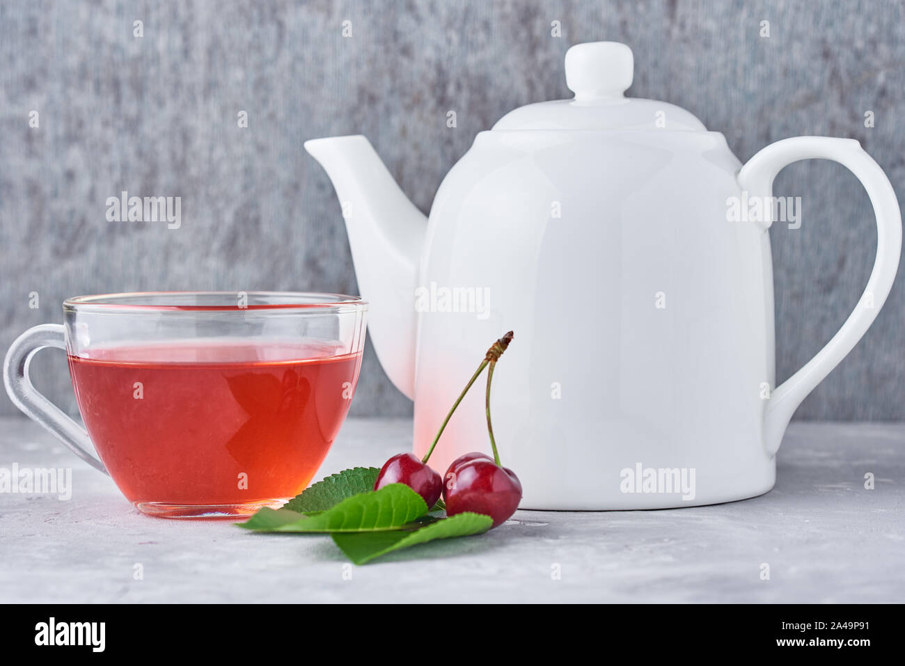 White teapot, herbal tea in glass cup and fresh cherry on gray