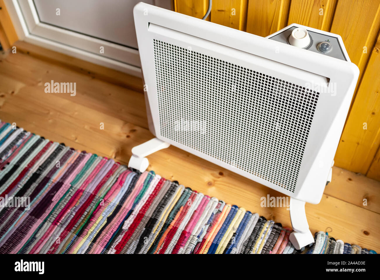 White electric radiator with a thermostat is placed on the floor covered with a colored rug, next to a wall and door, in a wooden country house. Stock Photo