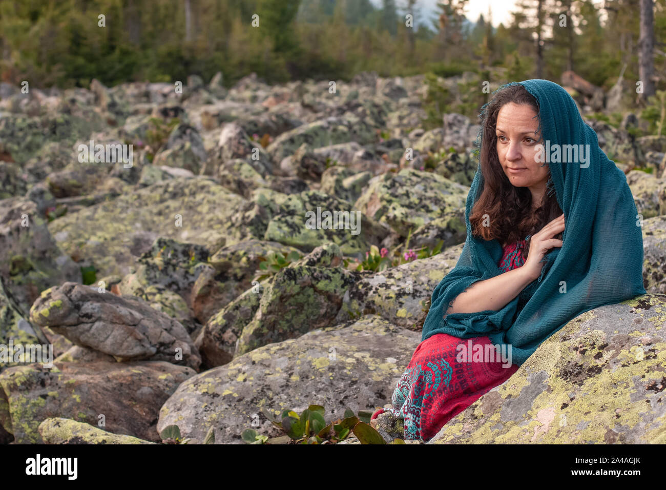 A girl with a scarf on her head sits by the large stones in the rock river. European appearance, dark hair. Red dress. On old stones, moss. Selective Stock Photo