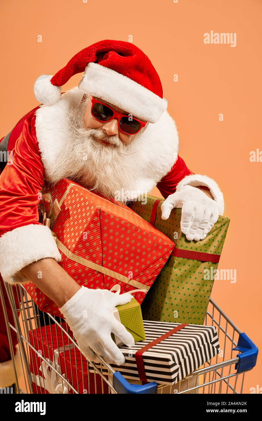 Cheerful Aged Man In Red Traditional Santa Costume Sunglasses And White Gloves Leaning On Shopping Trolley Filled With Colorful Gift Boxes Isolated On Orange Background Studio Shot Stock Photo Alamy