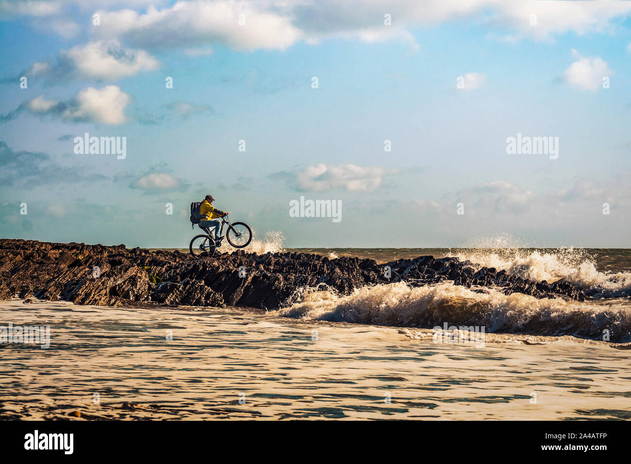 Cycling Ireland. Recreational cyclist with mountain bike and backpack trying to ride at sea rocks on the edge of wavy sea. Stock Photo