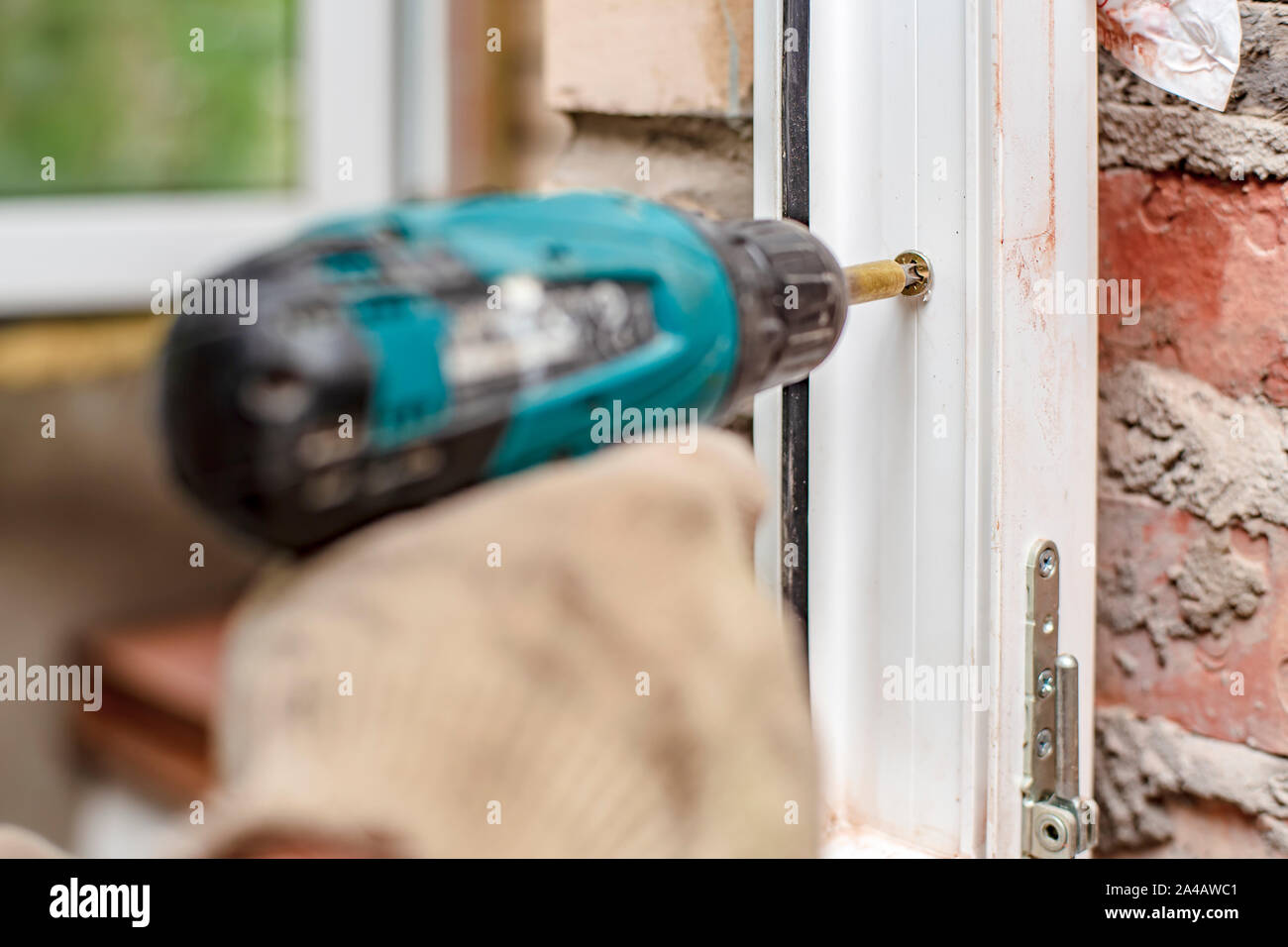 Male hands using an electric screwdriver and screws are going to fasten a PVC window frame to a brick wall. Close-up. Stock Photo