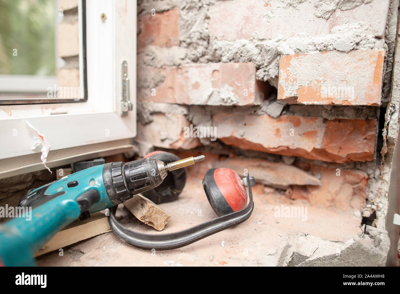 Noise-proof working headphones and an electric screwdriver are lying on a brick wall near a PVC window. Process of installing metal-plastic constructi Stock Photo