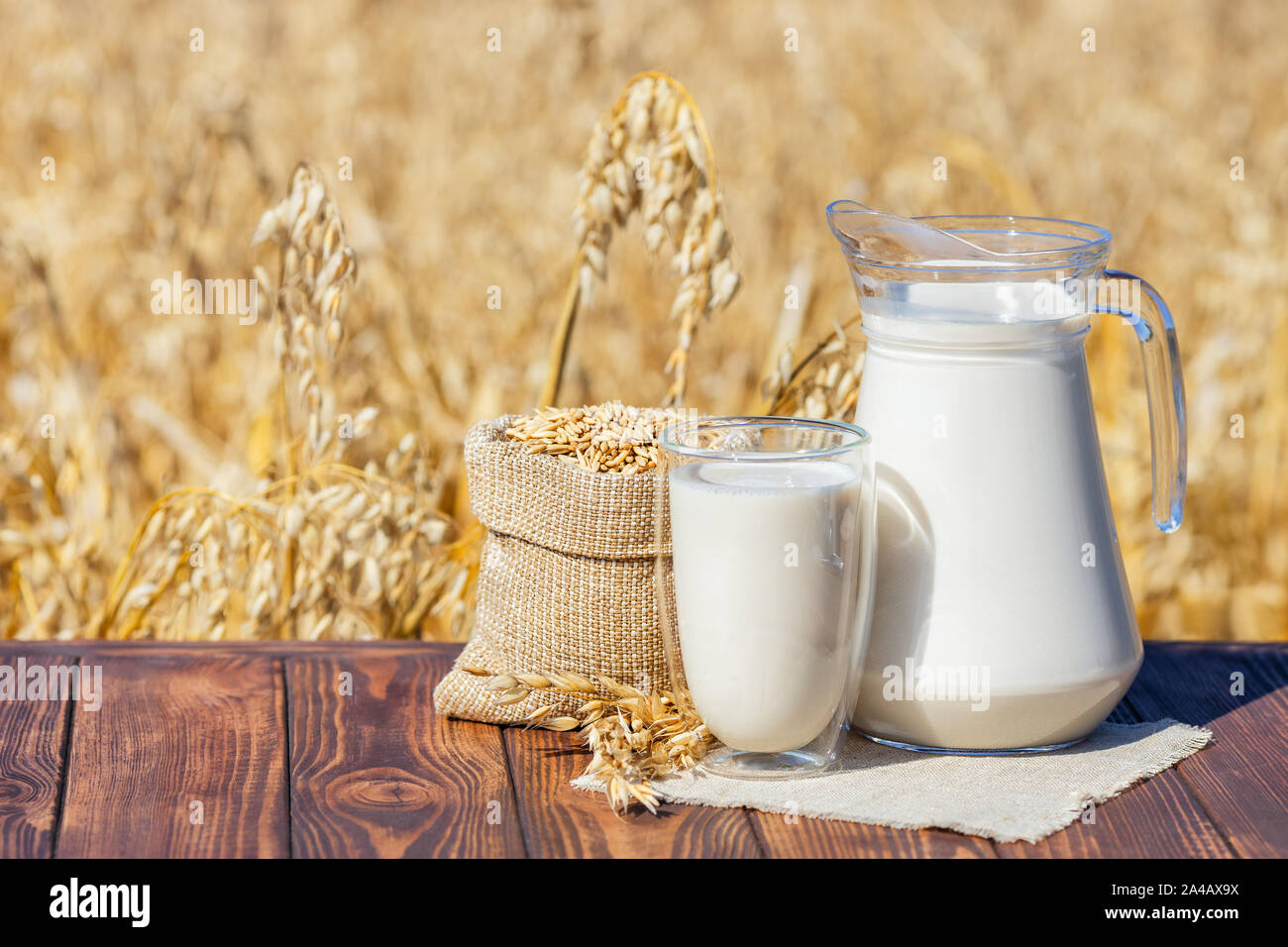 vegan oat milk in glass and jug with sack of grains on table over against ripe cereal field Stock Photo