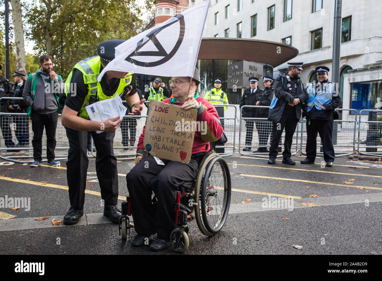 London, UK. 13 October, 2019. A police officer serves a notice under Section 14 of the Public Order Act 1986 to a disabled climate activist from Extinction Rebellion during a protest outside New Scotland Yard against tactics employed by police officers which impinge on the right to protest of disabled activists, including the confiscation of wheelchair ramps, accessible toilets and tents. Credit: Mark Kerrison/Alamy Live News Stock Photo