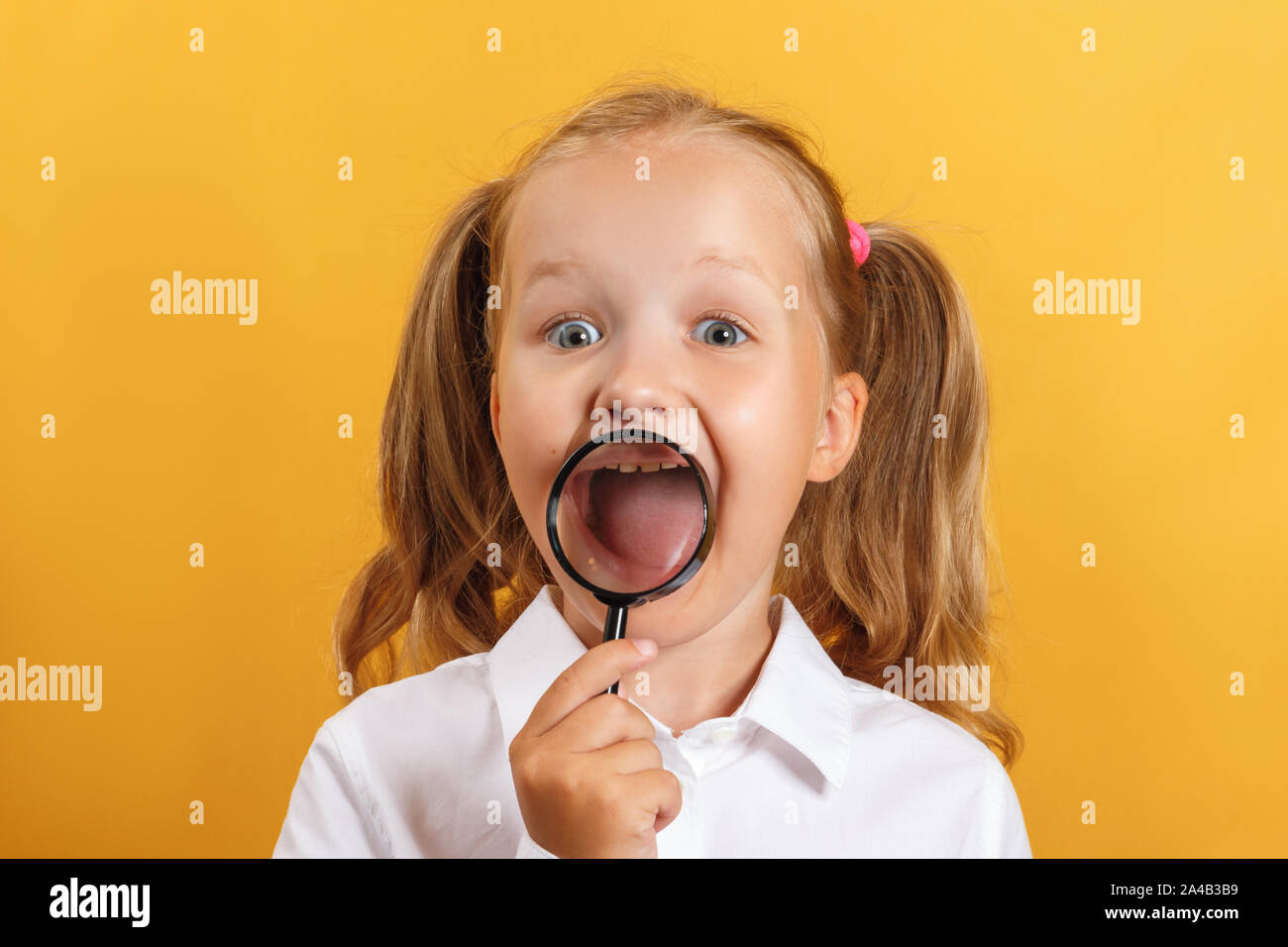 Cheerful schoolgirl little girl shows teeth and smiles through a magnifying glass. A child on a yellow background. Stock Photo