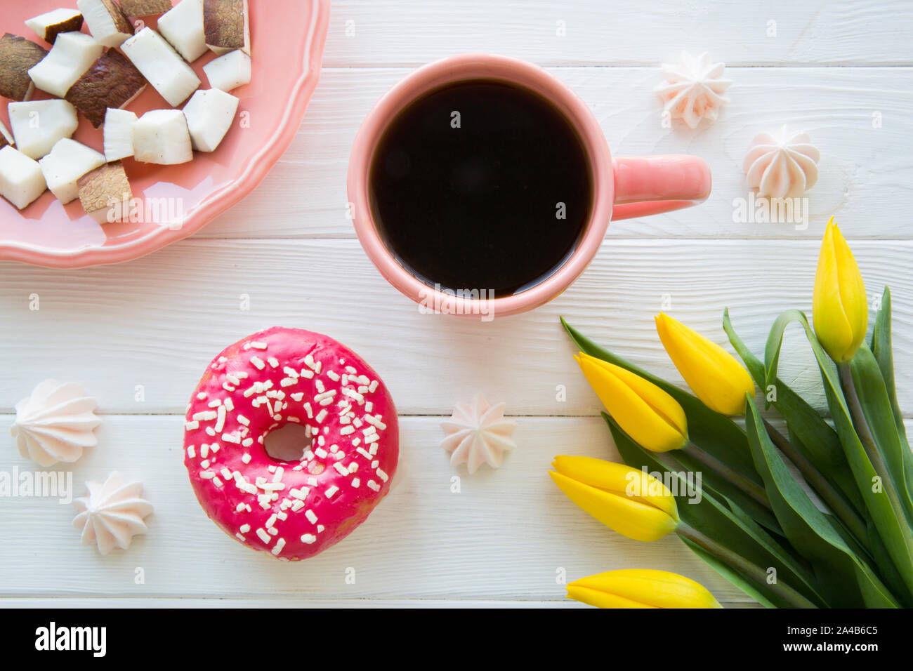 Sweet breakfast. Pieces of coconut, morning black coffee and pink glazed doughnut. Yeelow tulip decoration on white wooden background. Stock Photo