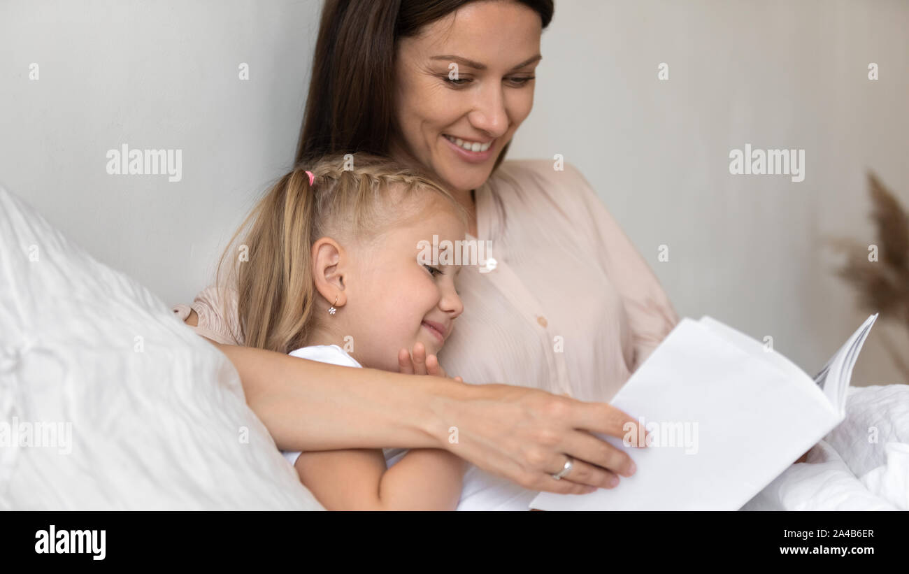 Smiling mother reading book to cute little daughter in bed Stock Photo
