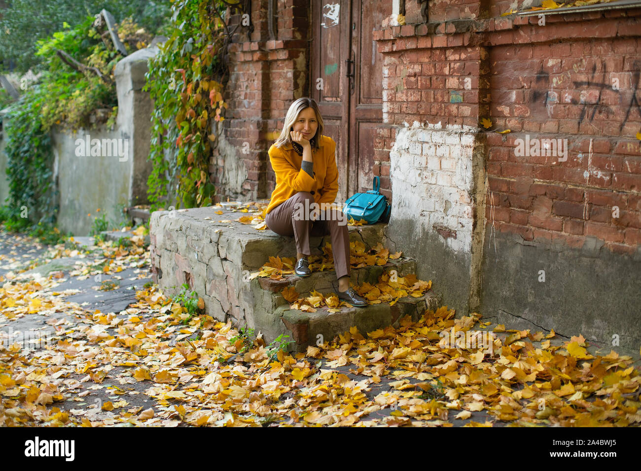 Attractive blonde woman in a yellow jacket poses in an autumn street with red and yellow foliage. Stock Photo