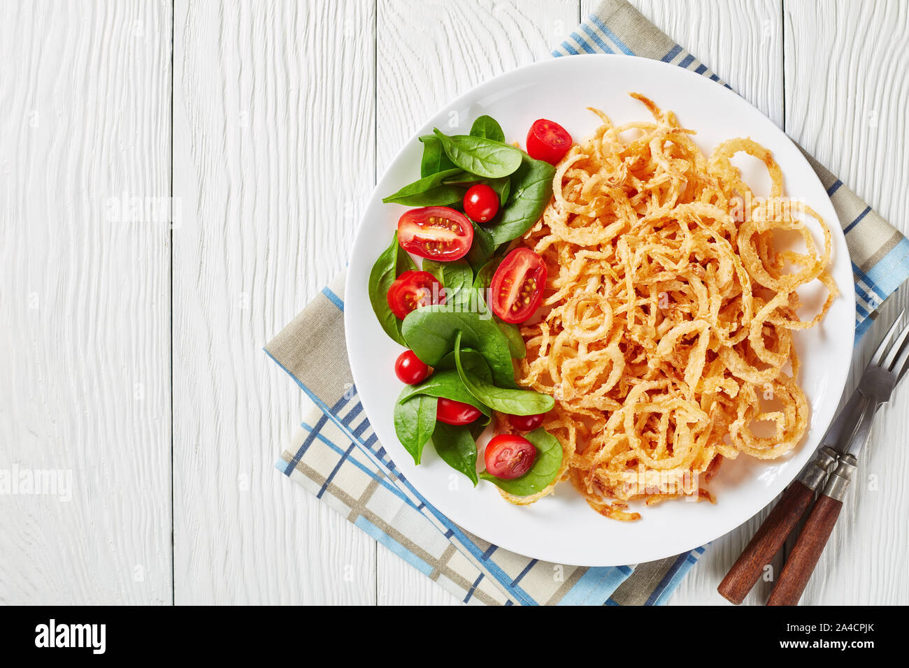 close-up of french crispy fried onion rings and strings with crunchy coating served with fresh spinach and tomato salad on a white plate on a wooden t Stock Photo