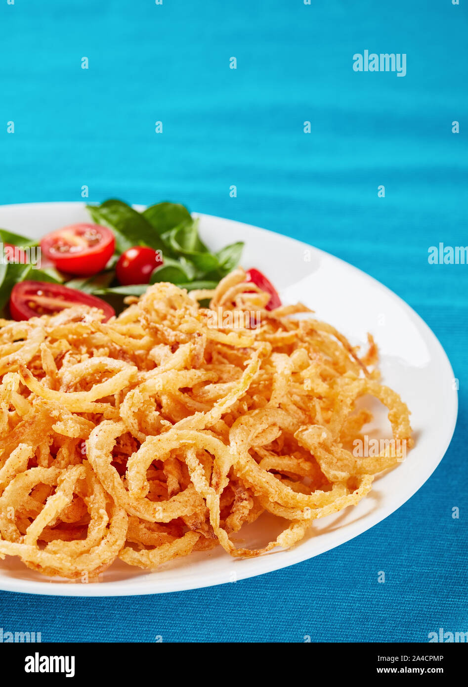 crispy fried onions with fresh spinach and tomato salad on a white plate on a textile table mat, vertical view Stock Photo