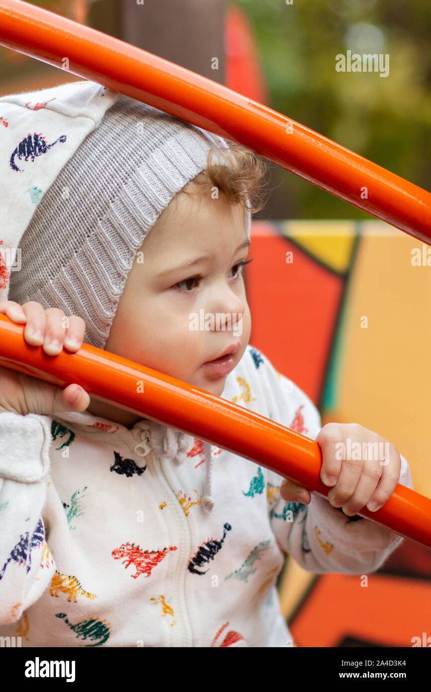 Baby Girl Boy In A Hoodie And Knitted Hat Looks Sideways Through A Red Metal Fence Caucasian Child On A Walk Holds On To The Railing Stock Photo Alamy