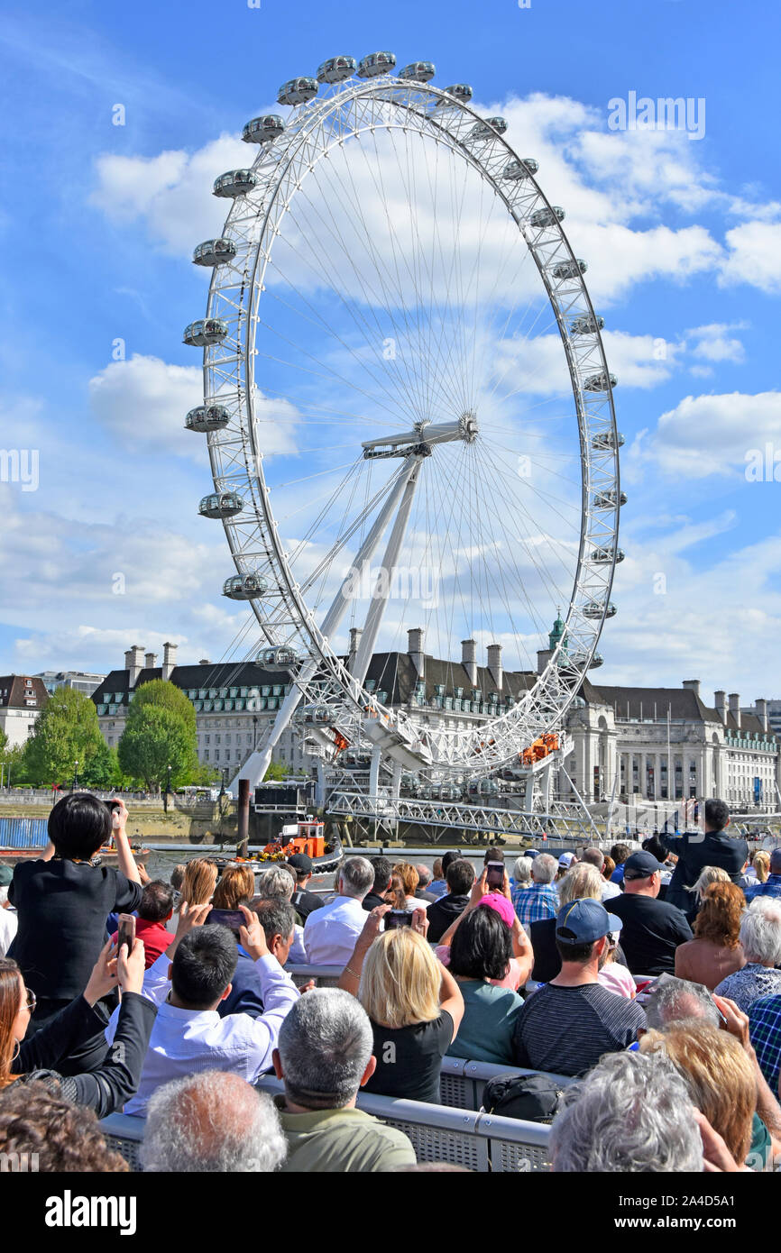 Back view group of people crowded River Thames sightseeing tour boat tourists taking mobile phone photo of iconic London Eye Ferris wheel England UK Stock Photo