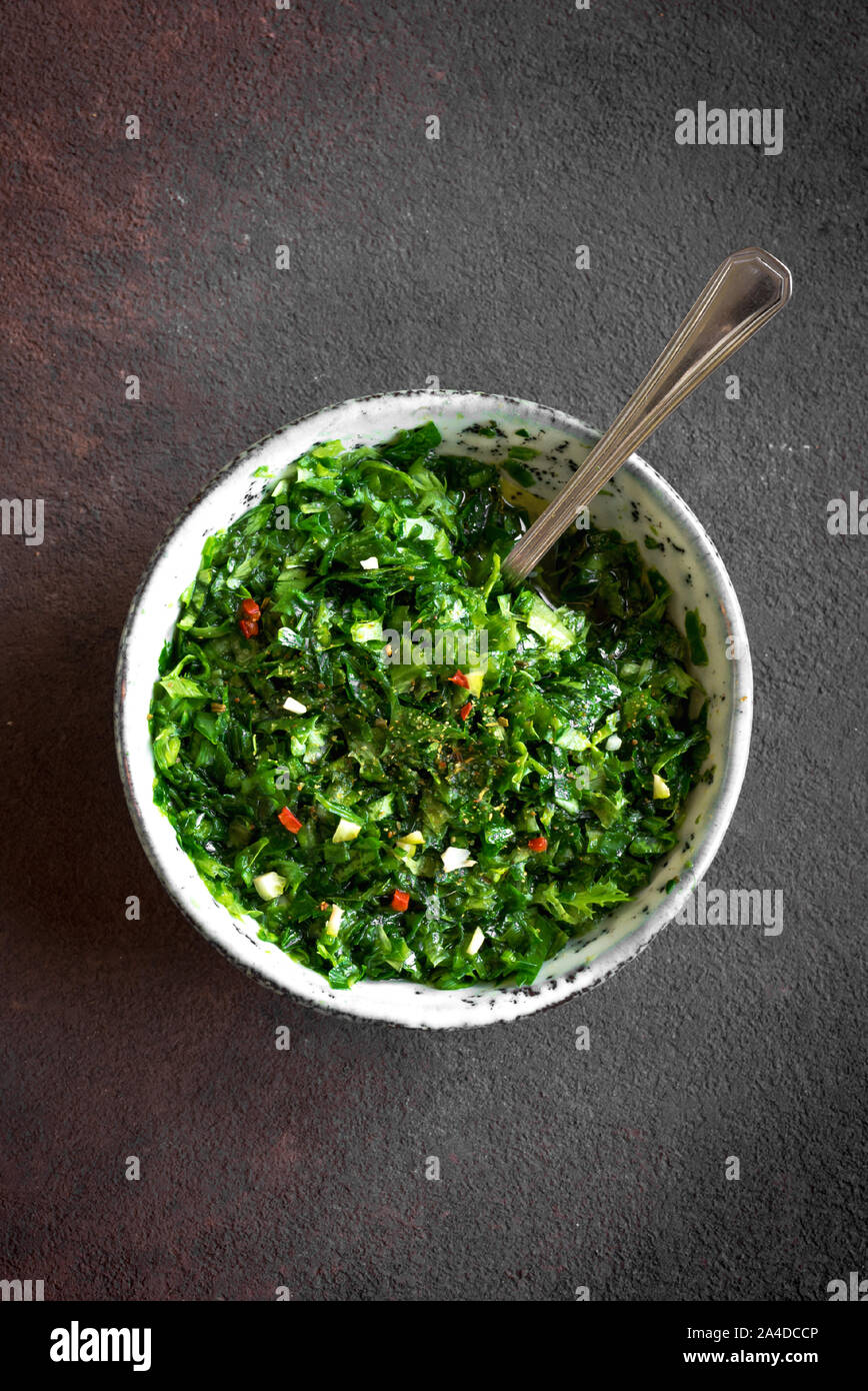 Homemade chopped green Chimichurri or Chimmichurri sauce made of parsley, garlic, oregano, hot pepper, olive oil, vinegar, on rustic table, close up. Stock Photo