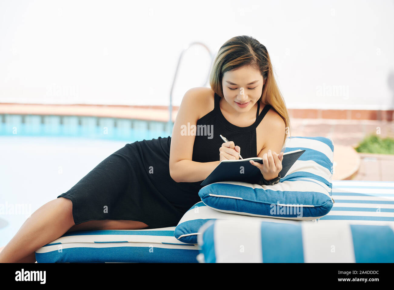 Beautiful smiling young woman in black bodycon dress lying on chaise-longe by the pool and filling gratitude journal Stock Photo