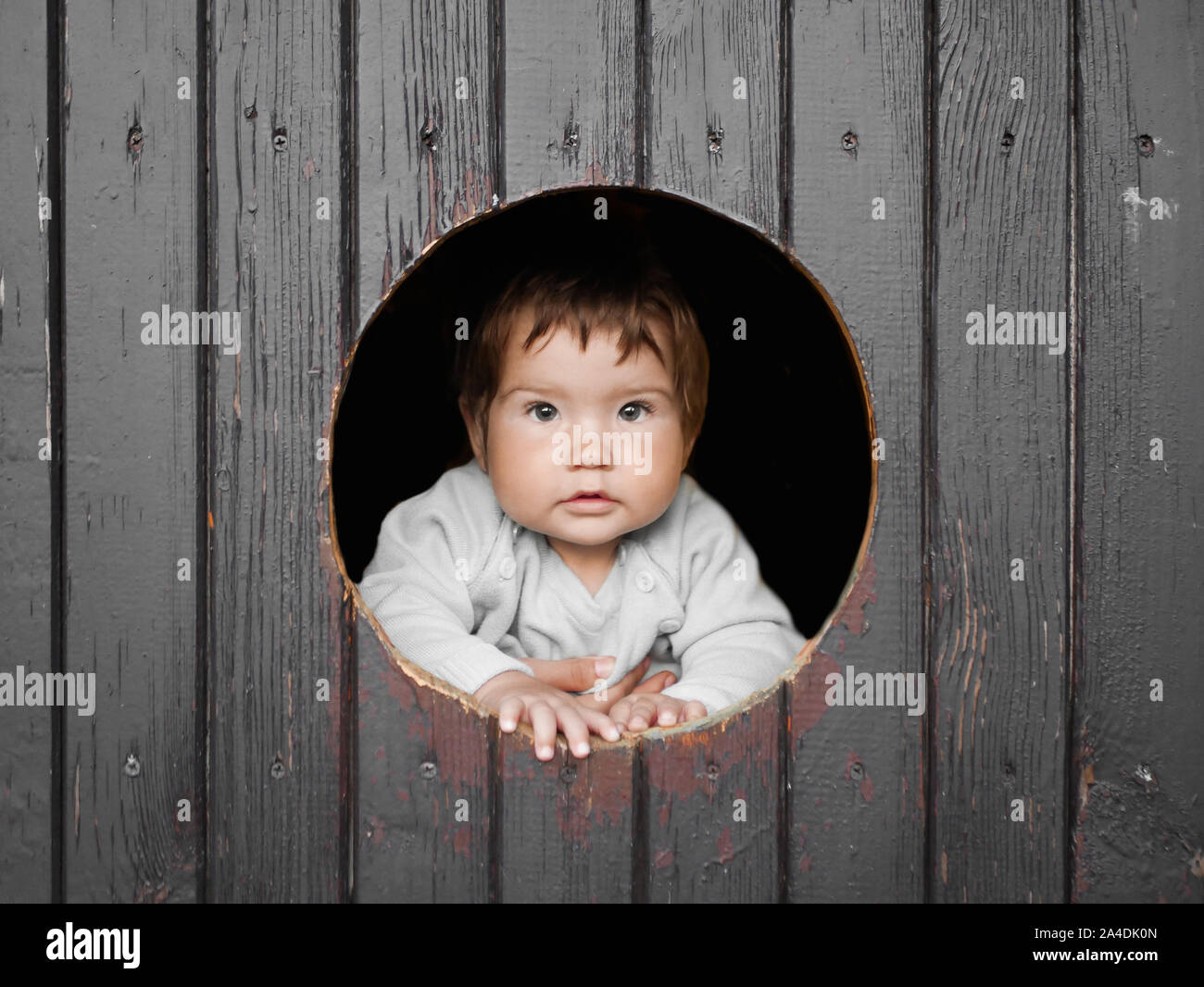 child looks out of a round wooden window and smiles. The boy climbed out of the round window and smiles pretty straight into the frame. Cheerful Stock Photo