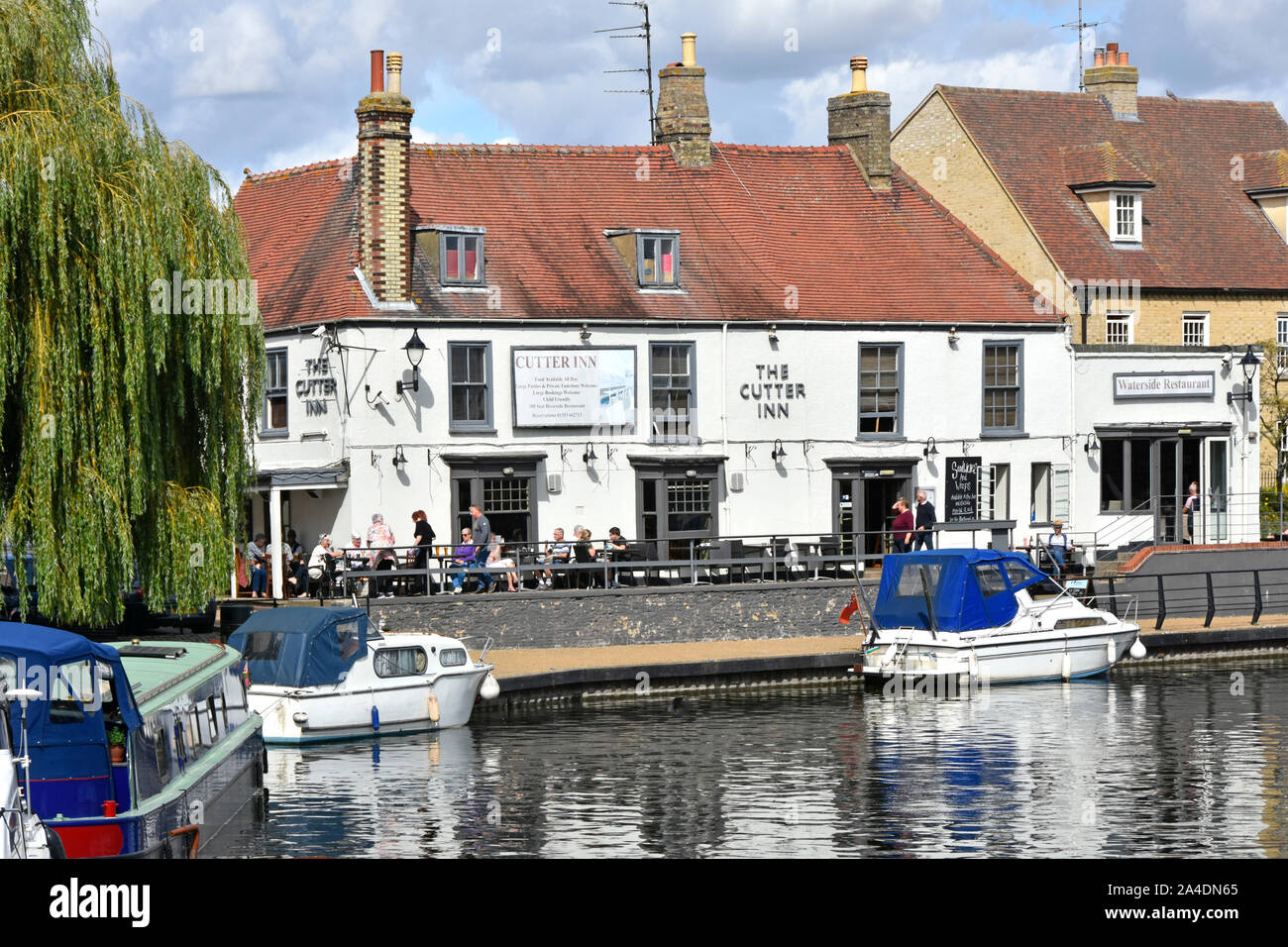 Lunchtime summer scene people outside The Cutter Inn pub & Waterside Restaurant river Great Ouse  boats beside towpath Ely Cambridgeshire England UK Stock Photo