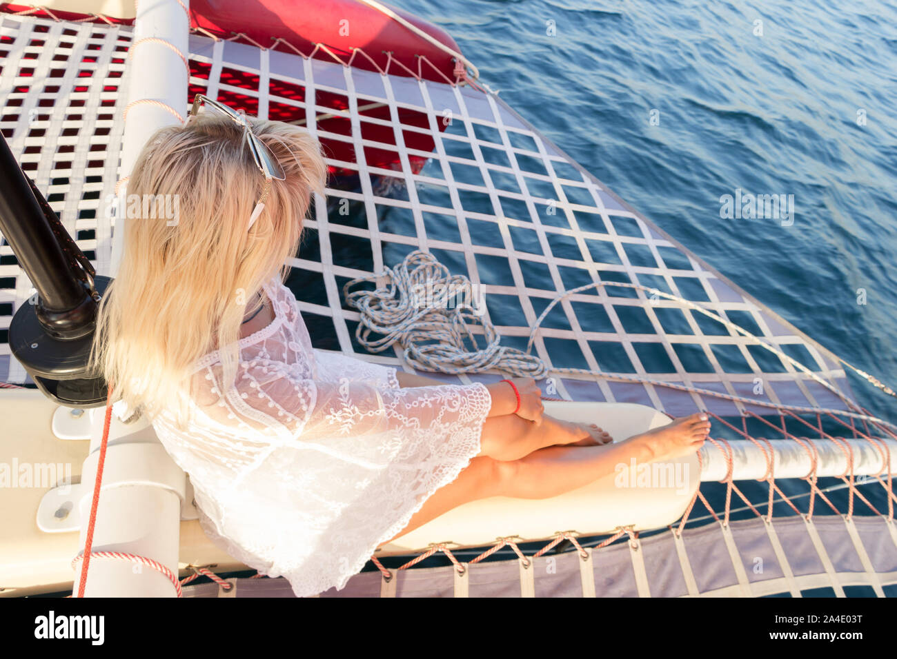 Beautiful young fair-haired woman dressed in a swimsuit and white pareo sitting on a yacht bow during sea journey. Side view. Selective focus on hair Stock Photo