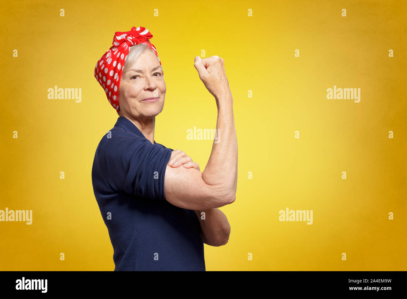 Rosie Riveter concept: proud elderly woman with red headscarf, flexing her muscles, copy space, yellow background Stock Photo