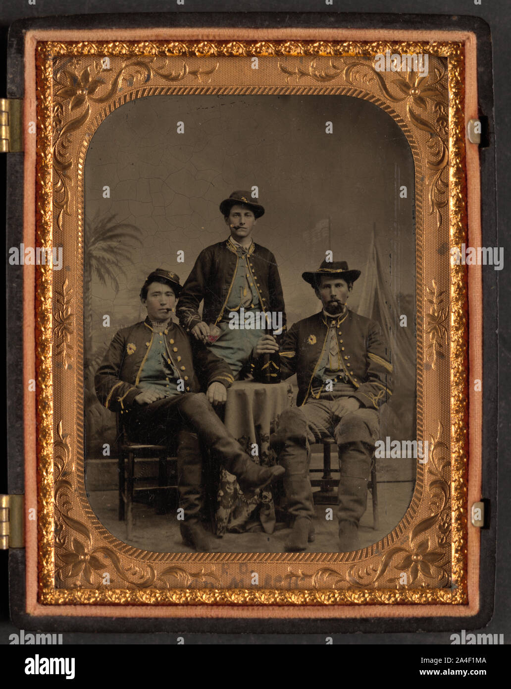 Three unidentified soldiers from the 75th New York Infantry Regiment who re-enlisted in Hancock's First Veterans Corps drinking wine and smoking cigars in front of painted backdrop showing palm tree and tents Stock Photo