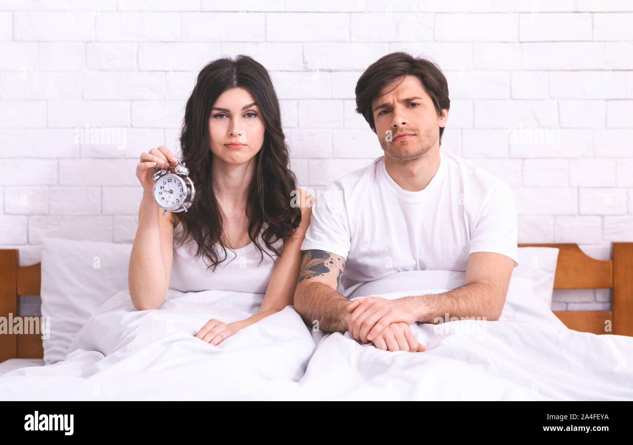 Sleepy couple sitting in bed with alarm clock in morning Stock Photo