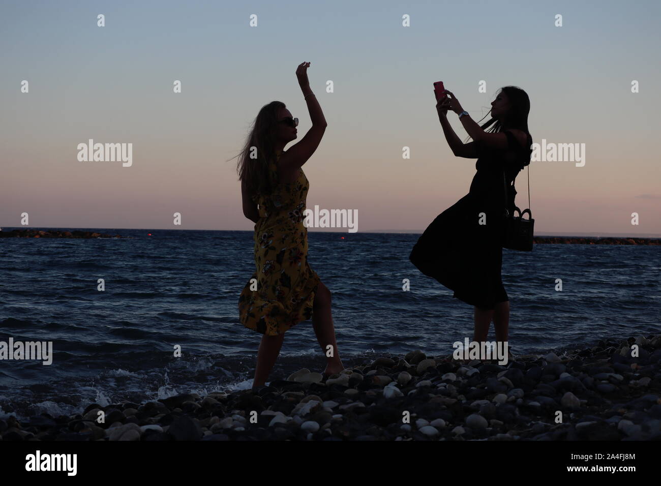 Limassol/Cyprus - August 15, 2019: Two young girls pose for pictures using smart phone camera on windy beach at sunset Stock Photo