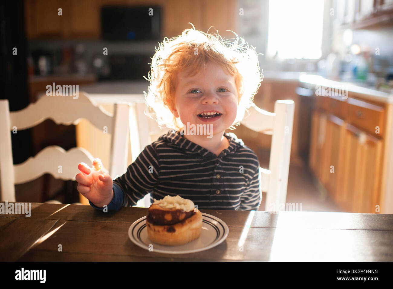 Toddler boy sitting at table enjoying a cinnamon roll at home Stock Photo