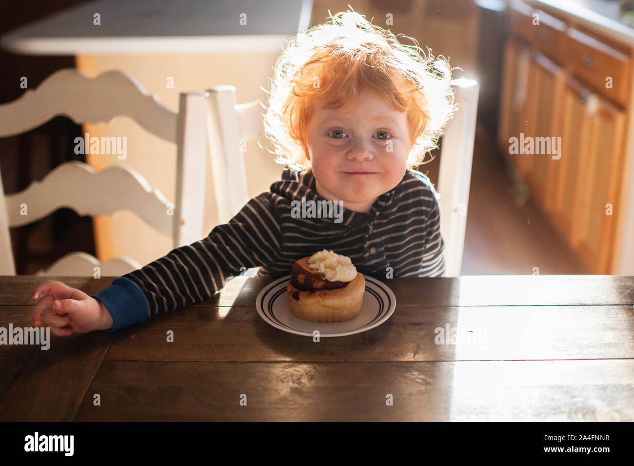 Toddler boy sitting at table enjoying a cinnamon roll at home Stock Photo