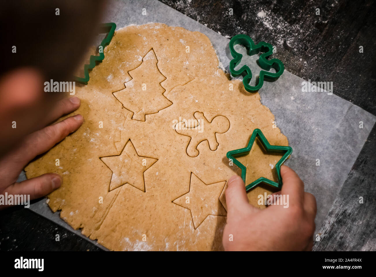 male hands cut out gingerbread cookie in the form of a snowflake, Christmas tree, a man from raw dough on parchment baking paper on a dark background Stock Photo