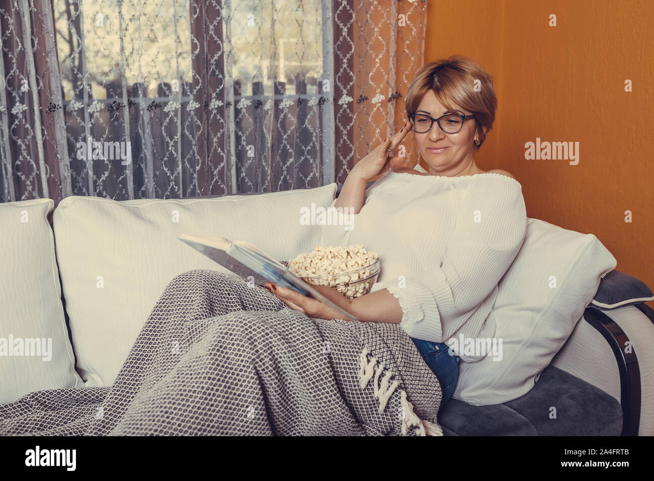 Middle aged woman reading book and eating popcorn while sitting on the couch at home Stock Photo