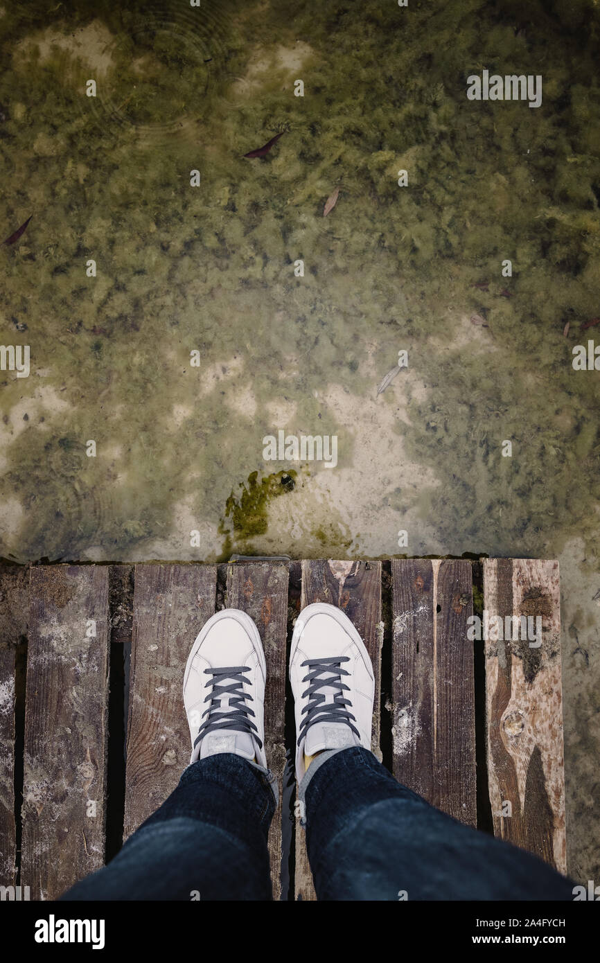top view of female legs in white sneakers and jeans on the background of an old dirty autumn lake-river and a wooden platform. Copy space Stock Photo