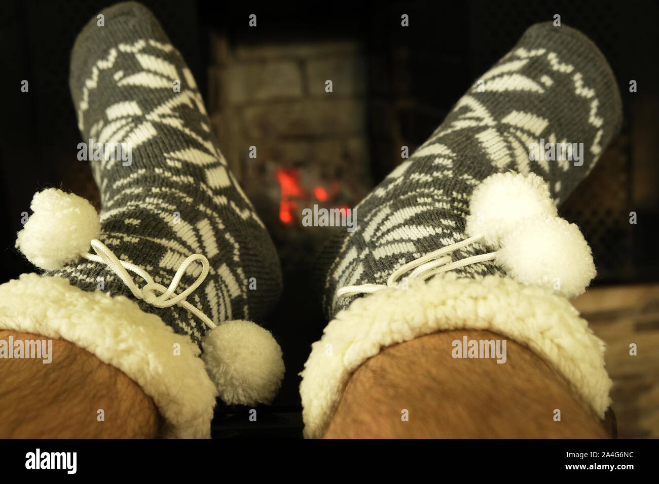 male hairy feet in woolen warm socks with ornament and white pom poms near fireplace in winter evening, concept of relaxing cozy New Year night Stock Photo