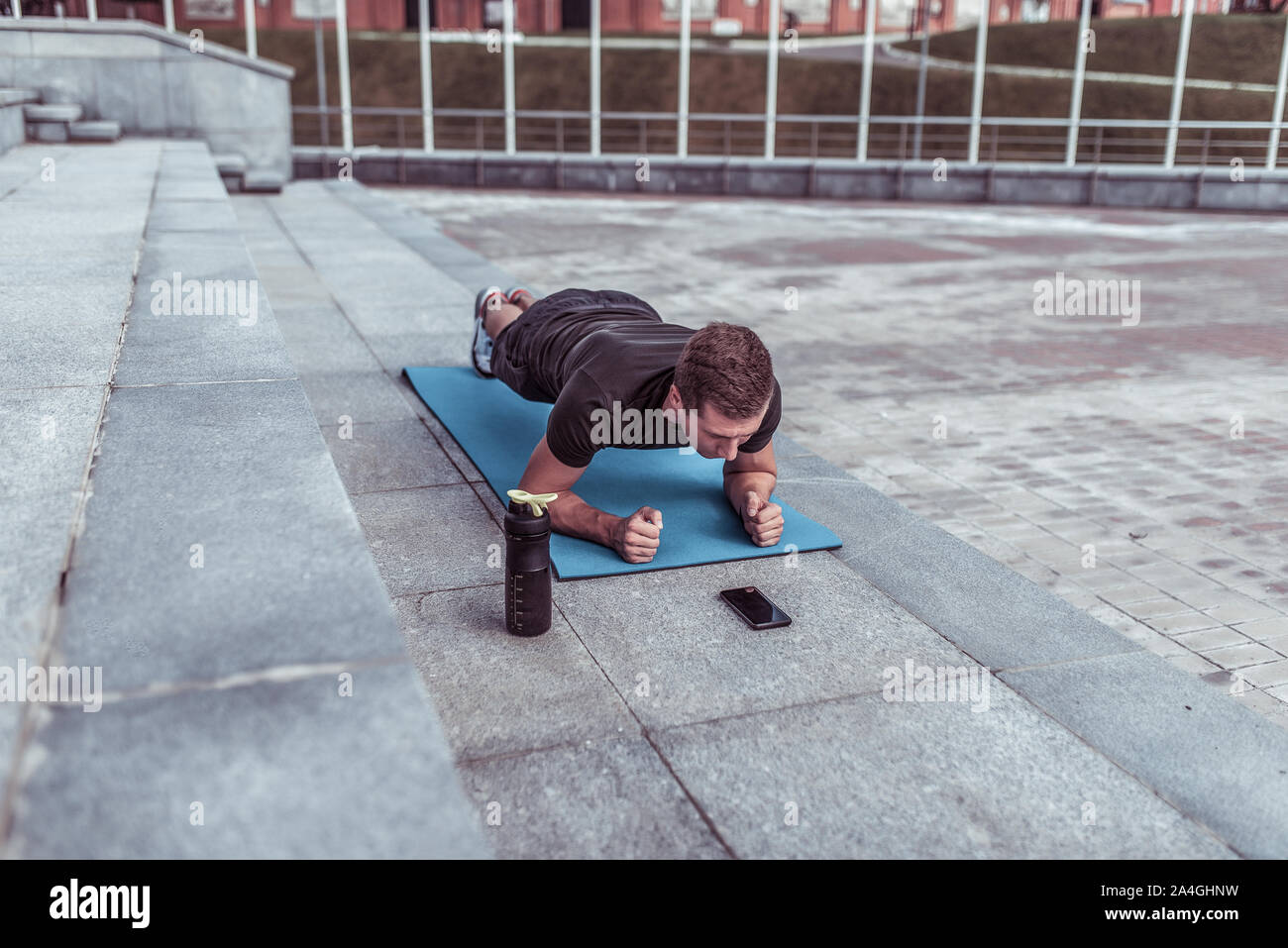 athlete man in summer in city, stands on bar, trains abdominal muscles and press, yoga mat, phone application in smartphone online timer, shaker with Stock Photo