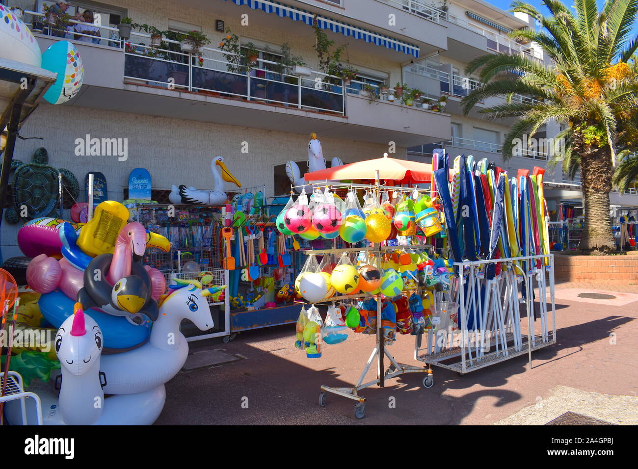 French shop on the promenade selling pool and beach inflatables, games, sets and toys. Palm tree and coastal residential building at the background. Stock Photo