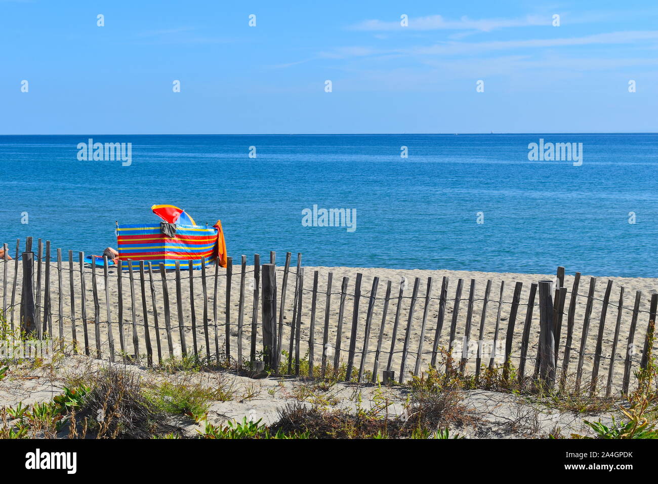 Colourful parasol and striped windbreak behind wooden fence on a miles long sandy beach in south of France. Calm blue sea meets the bright sky Stock Photo