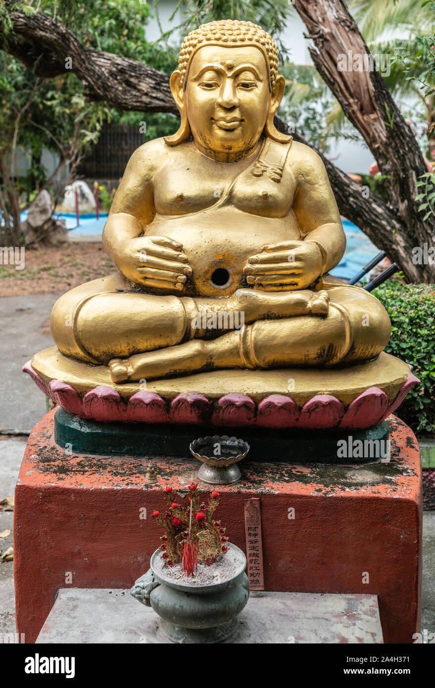 Bang Saen, Thailand - March 16, 2019: Wang Saensuk Buddhist Monastery. Gilded happy Buddha statue in garden, sitting on lotus, incense and flowers in Stock Photo