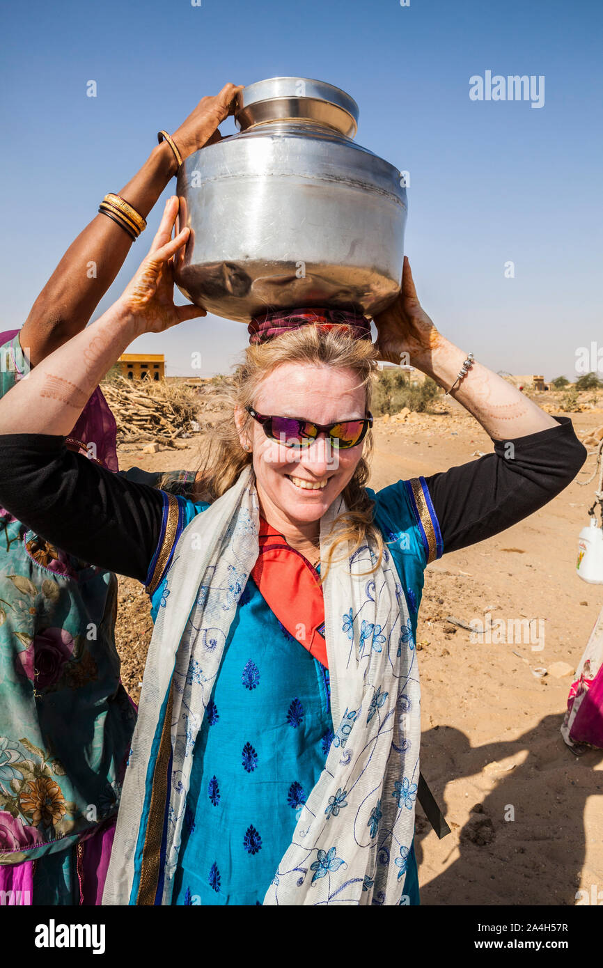A western tourist trying to carry a water jug on her head in a tiny village (Kanoi) in the Thar Desert, Rajasthan, India. Stock Photo